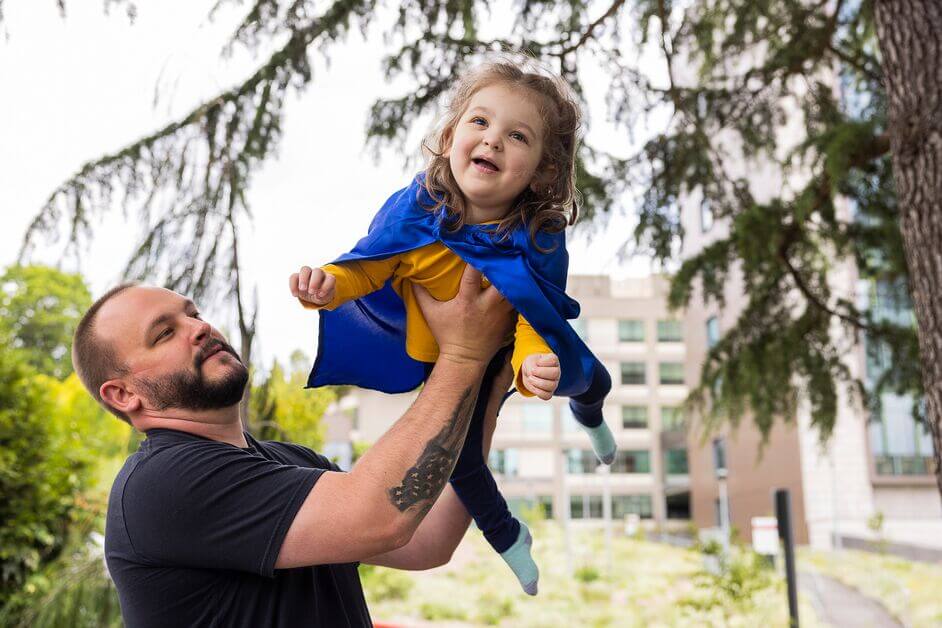 Little girl in yellow shirt and blue cape being lifted into the air by her father